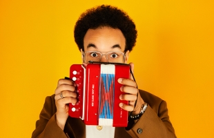 A close up shot of Théo Ould, he is pictured against a bright yellow background, holding up a small bright red and blue mini accordion, which covers the lower part of his face. He is wearing a glasses and he his eyes are open wide giving a surprised expression.