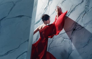 A lady wearing a dramatic red dress, with her back to the camera, holds her arm up against a marbled wall