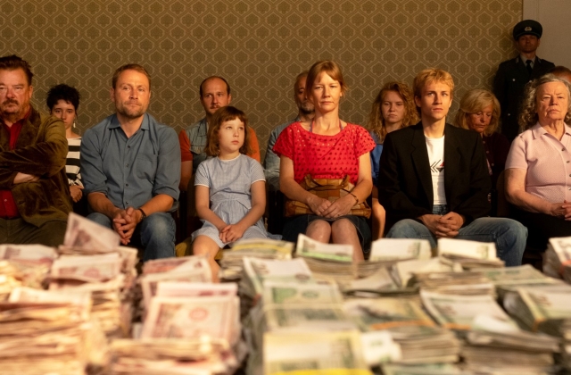 A selection of people, dressed in 80s/90s outfits, sit with a table covered in cash, wrapped in elastic bands