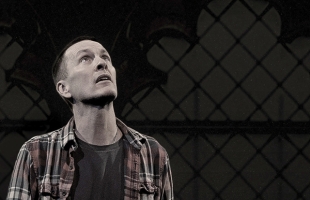 A man in a plaid shirt and jeans stands atop a wooden chair on a dark stage with his arms outstretched. Beside him, a framed projection screen displays a religious scene with a crucifix and figures in a church.