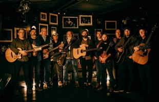 The full band of White Horse Guitar Club, stood in a dimly lit room holding their instruments