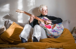 Wallis Bird, a woman with short light hair, sitting on a bed or sofa, playing an acoustic guitar decorated with colourful paint splashes. Sunlight streams across the wall and cushions behind her, creating soft shadows, while she looks down at the guitar as she plays.