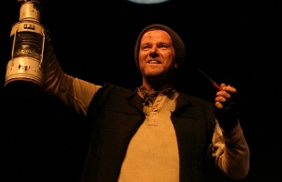 The photo depicts Aidan Dooley as Tom Crean. A man stands towards the camera, the moon is above him, one arm is raised holding up a lamp, the other arm is half raised holding a pipe. He is dressed in a hat and tunic.