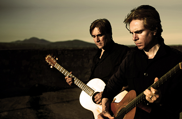 Brenan and Declan Murphy holding acoustic guitars and looking down pensively, in what appears to be a rural landscape at sunset