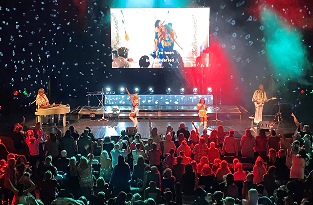 ABBA Forever performing for a packed audience in white outfits, with a projector screen behind them.