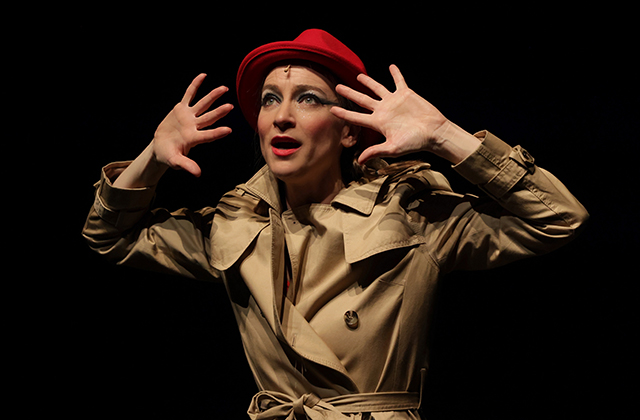 Sarah Louise You in An Evening Without Kate Bush, she is pictured on stage in a trench coat and a red trilby hat, her hands are up beside her head