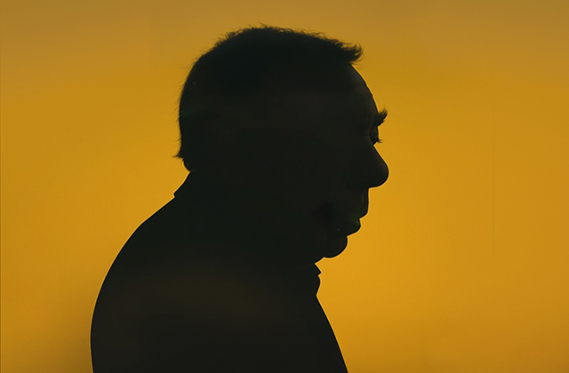 A side-profile silhouette of Andrew Lloyd Webber looking down against a bright, warm amber-yellow background. The lighting creates a dramatic, moody contrast, casting his head and shoulders entirely in shadow.