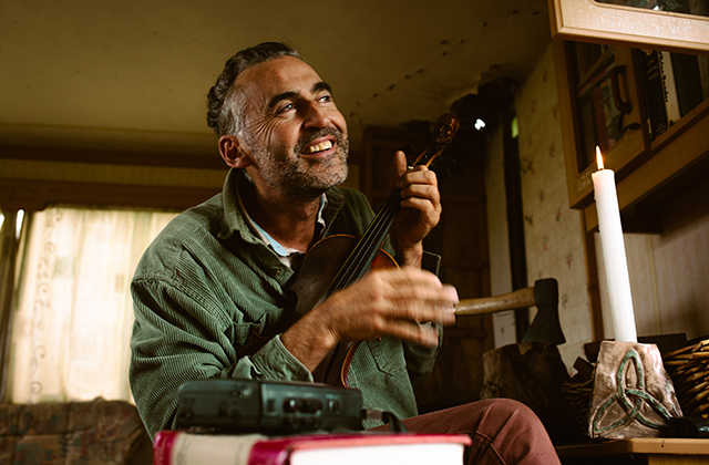 Aindrias de Staic, a smiling man with greying hair, sits in a warmly lit room, holding and playing a violin. A lit candle stands on the table beside him along with stacked books and a small record player, giving the cosy interior a rustic, intimate atmosphere. He looks upward as he plays, appearing relaxed and joyful.