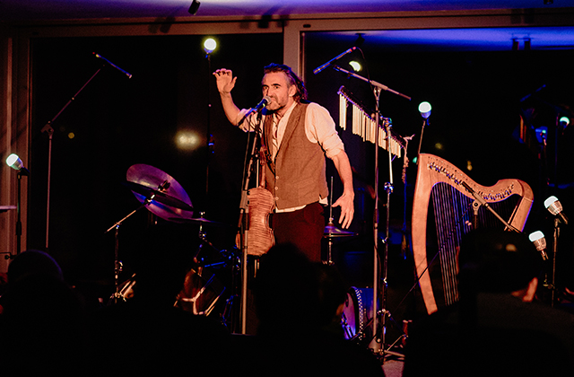 Aindrias de Staic stands on stage speaking into a microphone, raising one hand while performing. He wears a shirt and waistcoat, with a drum kit and a harp set up behind him. The stage is lit with colourful lights, and audience silhouettes are visible in the foreground.