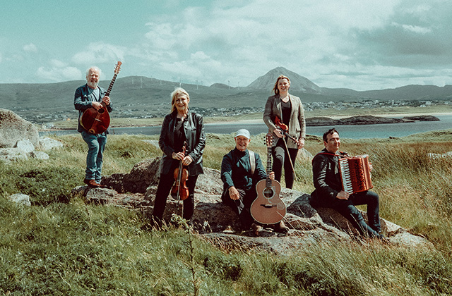 Altan poses outdoors on rocky grassland beside a coastal bay, holding traditional instruments including guitars, a violin, and an accordion, with mountains and a cloudy sky in the background.