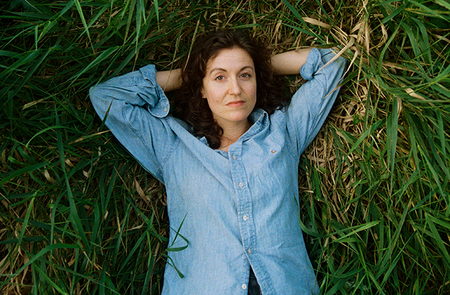 Tessa Rose Jackson lying on their back in long green grass, viewed from above. Both arms are bent with hands resting behind the head. The person wears a light blue, long-sleeved button-up shirt. Dark hair spreads slightly into the grass. The surrounding grass fills the entire frame, with a mix of fresh green blades and flattened straw-like patches creating a textured natural background.