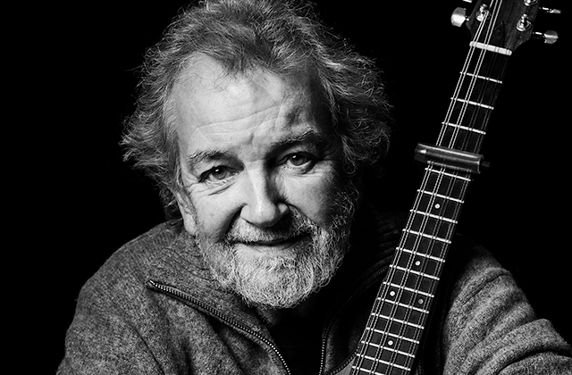 A black-and-white portrait of Andy Irvine who has a beard and tousled hair, smiling gently while holding a guitar close to his face.