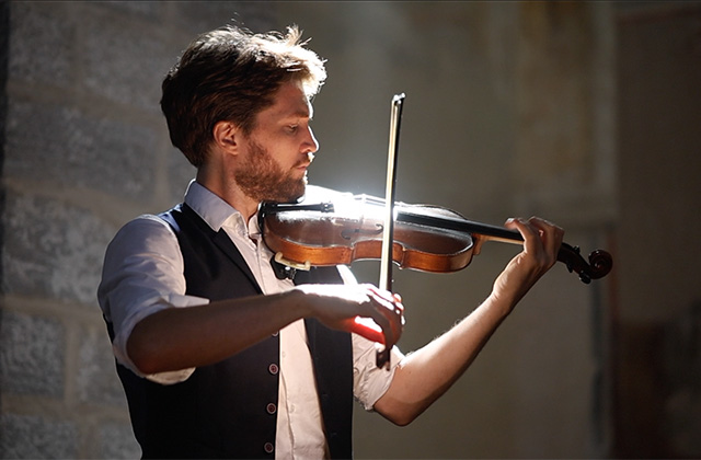 Anton Jablokob playing a violin under warm, dramatic lighting indoors, wearing a white shirt and dark vest while focusing on the performance