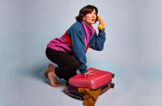 A photo of comedian Aoife Dunne wearing a pink, blue and orange fleece with brown trousers. She is crouched beside a red suitcase and holding a yellow telephone to her ear.