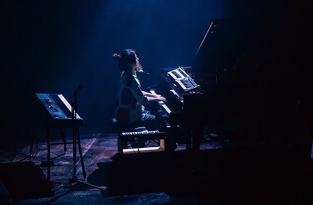 Belle Chen sits at a grand piano on a dark stage, lit by a cool blue spotlight. She plays with focused energy, surrounded by keyboards and microphones, creating a dramatic and atmospheric performance scene.