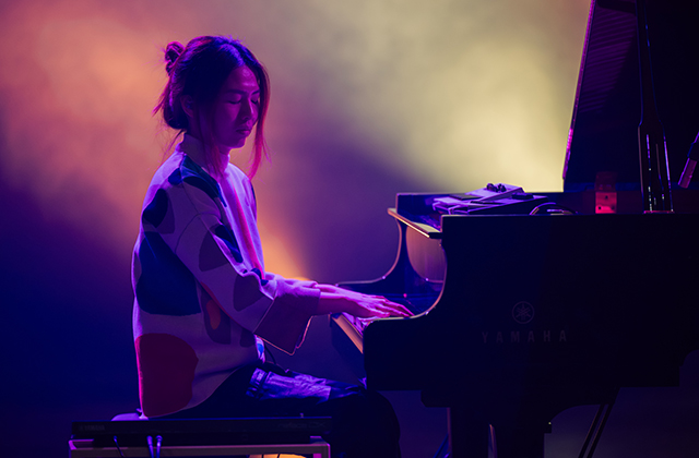 Belle Chen plays a Yamaha grand piano onstage, illuminated by warm, colourful lighting. She appears calm and concentrated, wearing a patterned top as soft hues of purple, yellow, and orange glow behind her.