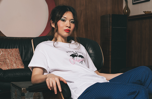Belle Chen sits relaxed in a black leather chair in a cozy, mid-century styled room. She wears a white graphic T-shirt and blue patterned pants, with her hair tied back and bold makeup. One arm rests on the chair’s armrest while she gazes confidently toward the camera.