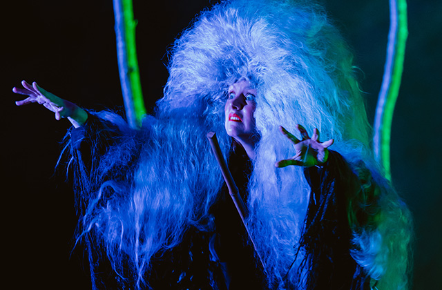 Bryony Kimmings is pictured in a scene from Bog Witch. She is in a flamboyant costume with a large fluffy headress on, her arms are reached out like a monster and she has a wild look on her face. The lighting illuminates her in blue & green hue.