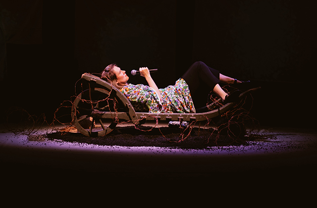 Bryony Kimmings is pictured in a scene from Bog Witch. She is holding a microphone, wearing a summer dress and is lying in a wooden carriage surrounded by red barbed wire.