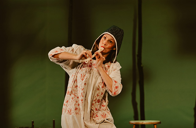 Bryony Kimmings is pictured in a scene from Bog Witch. She is wearing a medieval looking night dress and green hat, she is playing a recorder in a jester like manner