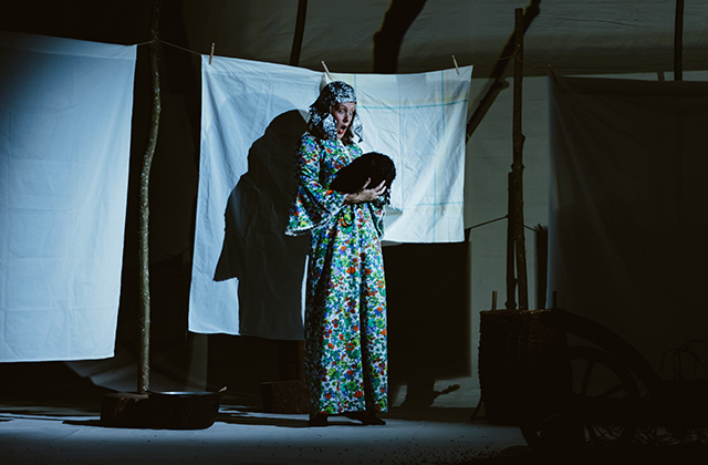 Bryony Kimmings is pictured in a scene from Bog Witch. She is standing wearing a metallic headdress, and long flowery dress,  she stands in front of a washing line and is looking surprised