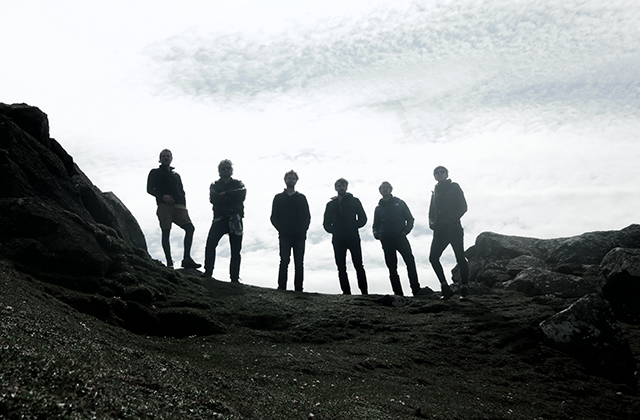 Silhouetted group of seven people standing along the edge of a rocky outcrop, viewed from below. Dark rock formations frame the foreground on both sides, while a bright, cloud-filled sky creates strong contrast behind the figures. The people are spaced evenly, facing forward, with minimal visible detail due to backlighting.