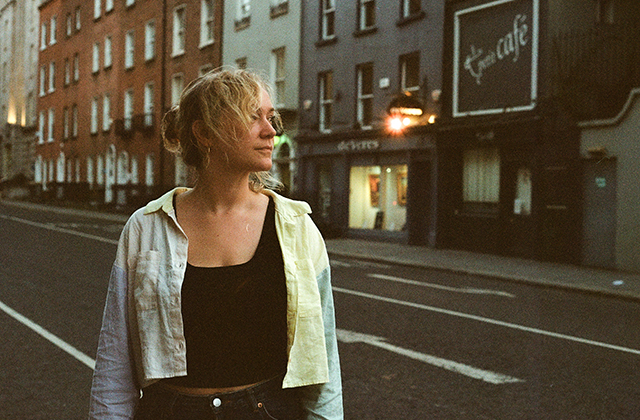 Person standing on a quiet urban street at dusk, photographed from the waist up. The face is obscured. The person wears a light jacket over a dark top. Behind them are brick buildings with shopfronts and signage, softly lit streetlights, and empty road markings, creating a subdued evening city atmosphere.