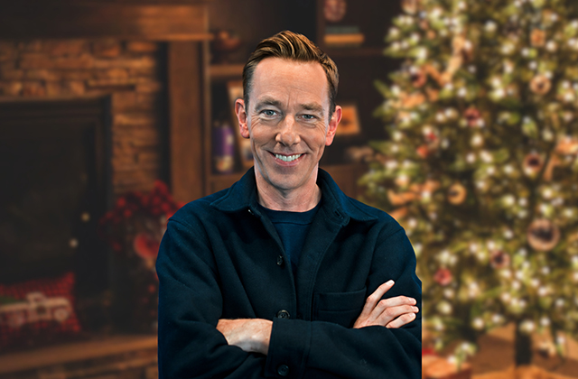 Ryan Tubridy smiles at the camera with his arms crossed in a medium shot. He wears a dark navy button-up jacket over a dark top. The background features a cozy, softly blurred indoor holiday scene, including a stone fireplace on the left and a decorated Christmas tree with lights and ornaments on the right.