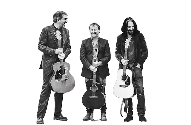 From left to right, Fiachna, Tom and Alan are standing together dressed in suit jackets holding their guitars, they are all smiling. The photo is black and white against a brilliant white background.