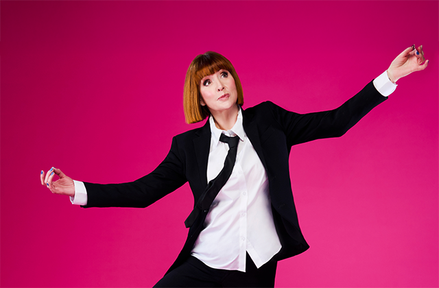 Cally Beaton, dressed in a black suit, with white shirt and black tie, holds her arms out with her fingers together in the classic