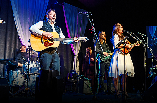 Cash Returns tribute band performing live on stage. JP Mac, portraying Johnny Cash, stands at the front left playing an acoustic guitar and singing, while Karen Martin, as June Carter Cash, stands to the right in a white dress playing a mandolin. A full band including drummer, bassist, and backing vocalist perform behind them, with colourful stage lighting and draped curtains enhancing the scene.
