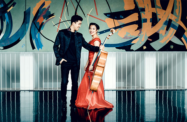 Bryan Cheng, wearing an all black suit and holding his cello out beside him, stands alongside Silvie Cheng dressed in a long red silk gown