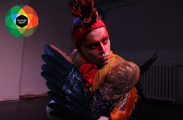 Production still of 'Chicken' - Eva O'Connor dressed as a cockerel looking over her right shoulder at the audience