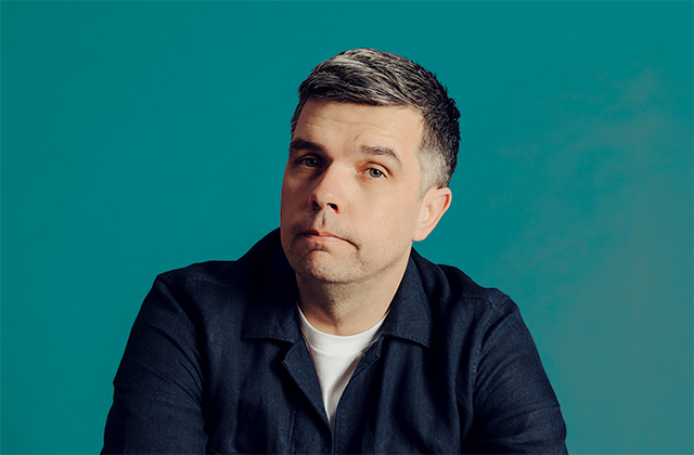 A studio portrait of a Chris Kent, a man with short dark hair and light stubble, wearing a dark jacket over a light shirt. He faces slightly to one side with a neutral expression. The background is teal with soft, even lighting.