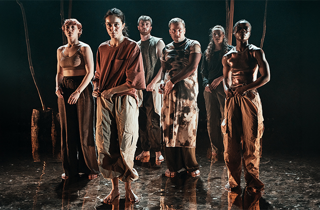 Six barefoot contemporary dancers stand in formation on a dimly lit stage, wearing loose earth-toned costumes, with dramatic lighting reflecting off the floor.