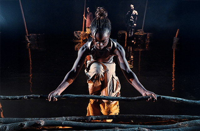 A young black female dancer, dressed in a gold top, and orange trousers bends down to pick up a large stick