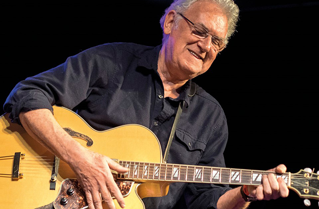Jim Mullen, an older man wearing glasses plays a hollow-body electric guitar on stage, smiling as he performs under stage lighting.