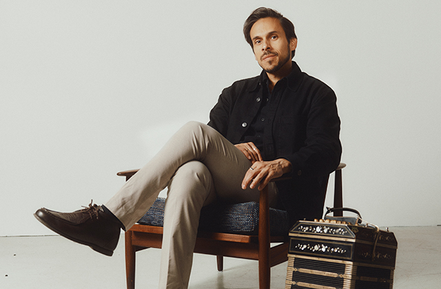 Man with dark hair and light stubble sits relaxed in a wooden armchair against a plain light background. He wears a black jacket, dark shirt, beige trousers, and brown leather shoes, with one leg crossed over the other. His expression is calm and thoughtful. A bandoneon (accordion-like instrument) rests on the floor beside him.