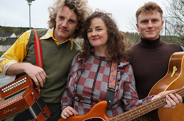 Three musicians standing closely together outdoors. On the left, a man with curly blonde hair holds an accordion, wearing a green sweater vest over a patterned yellow shirt. In the center, a woman with dark curly hair holds an electric bass guitar while wearing a grey and red patterned long-sleeve dress. On the right, a man with short light hair wearing a dark turtleneck holds an acoustic guitar.