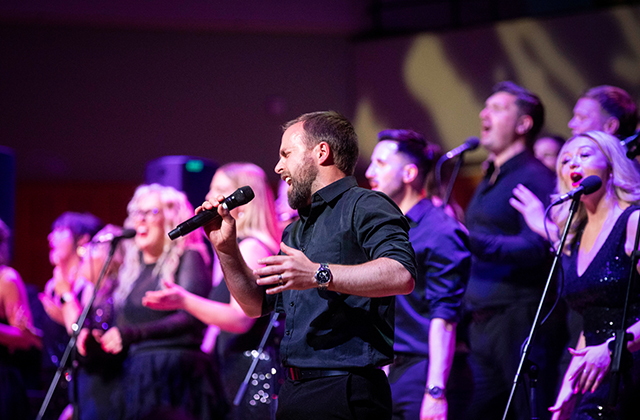 A group of singers, dressed in black, with microphones, singing passionately