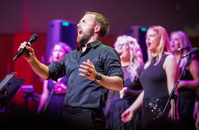 A bearded male vocalist in a black shirt sings passionately into a handheld microphone with his eyes closed and free hand raised. Behind him on stage, several female backing vocalists in black outfits sing along under vibrant purple and red stage lighting.