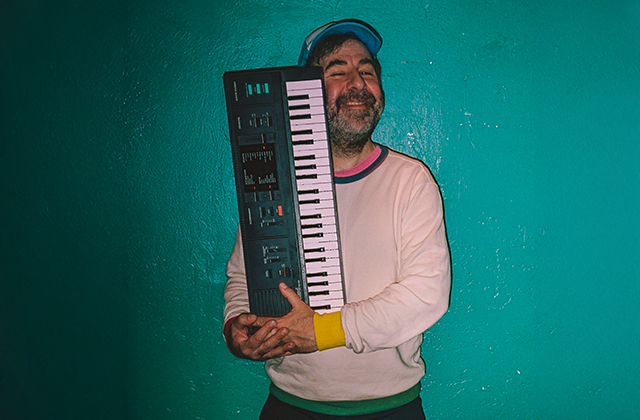 David O'Doherty stands wearing a cap & jumper. He is hugging his keyboard with a happy smile leaning against a green coloured wall.