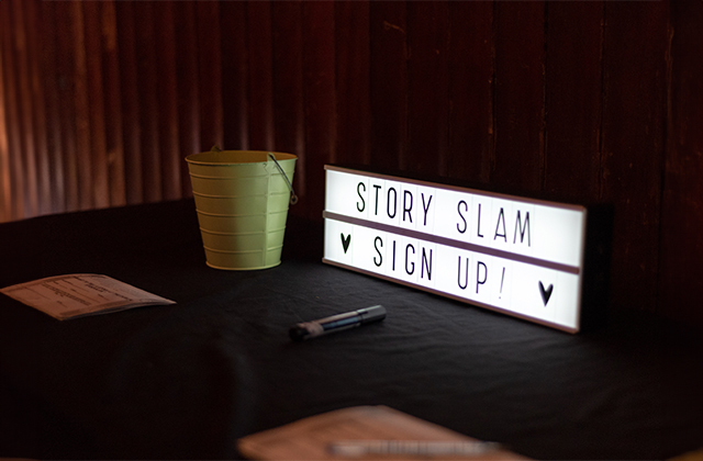 A light box with the words STORY SLAM on the top line, and on the bottom line SIGN UP! with heart emojis either side.