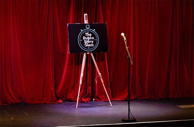Image of a stage, with a red curtain at the back. Centre stage, in the spotlight is an easel with the Dublin Story Slam logo being held up. A microphone stands in front of it.