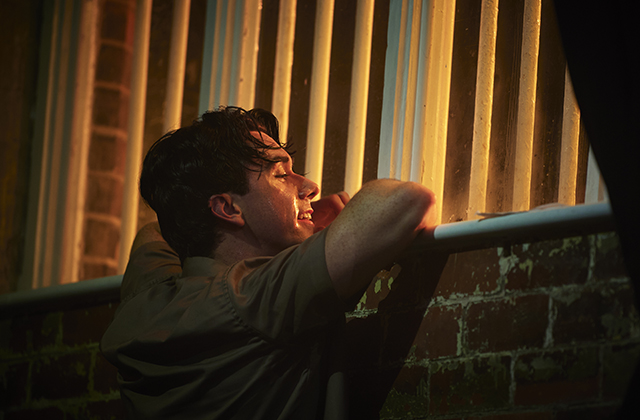 A man leans over a brick wall, looking out a window with vertical blinds