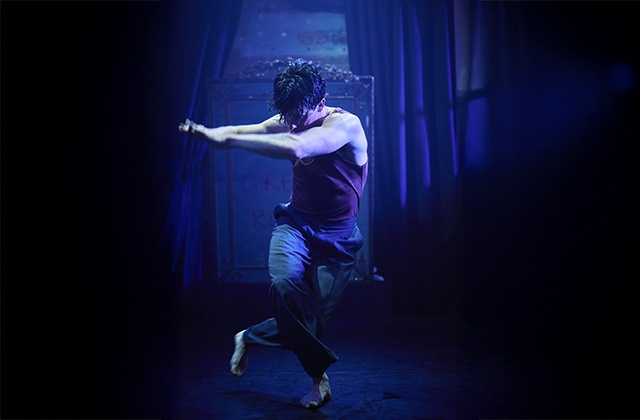 A male dancer, wearing a vest and trousers strikes a pose mid-dance centre stage, swathed in blue light