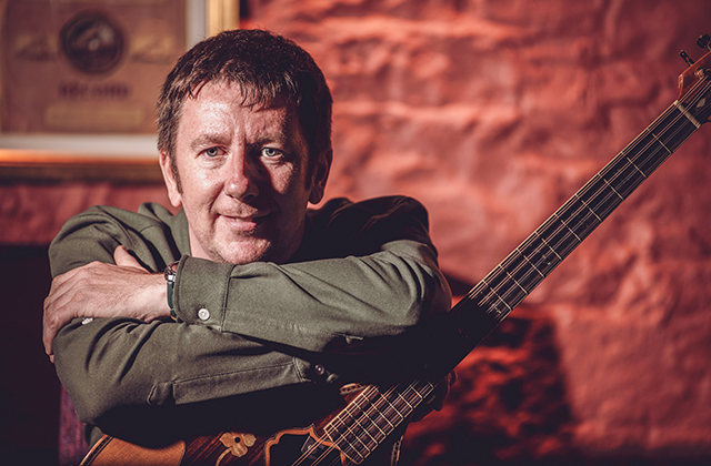 A man with short dark hair leans his arms on an acoustic guitar, posing in a warmly lit room with a stone wall behind him.