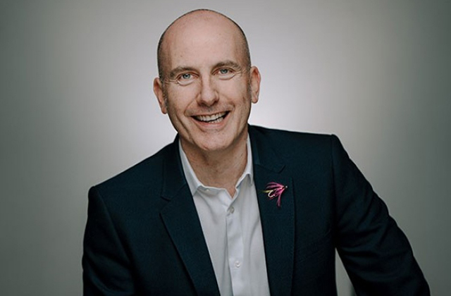 A portrait of David, he sits wearing a suit, he is smiling towards the camera