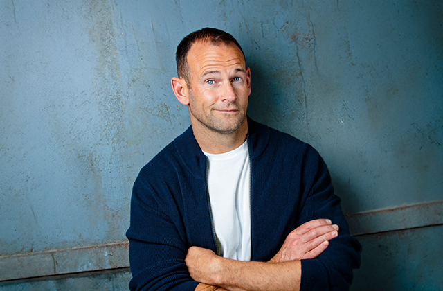 David Nihill with his arms crossed, smirking at the camera, with a blue wall behind him