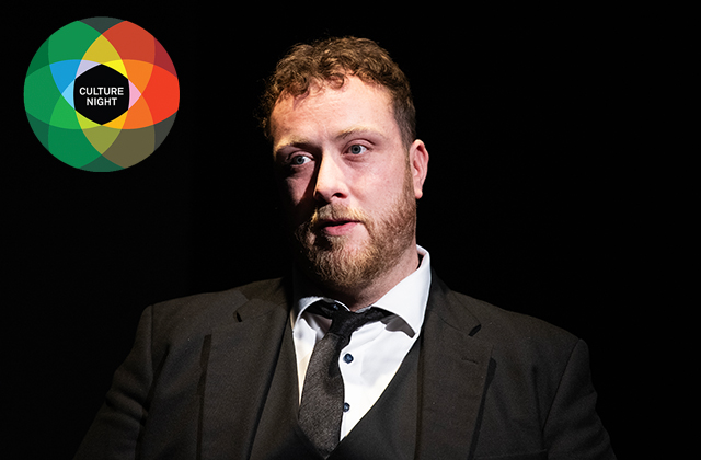 A man with short curly brown hair, a light beard, and a serious expression illuminated against a dark background. He wears a dark suit jacket, a matching waistcoat, a white collared shirt, and a dark tie. In the top left corner, a circular logo made of overlapping colored circles surrounds a black center with the text